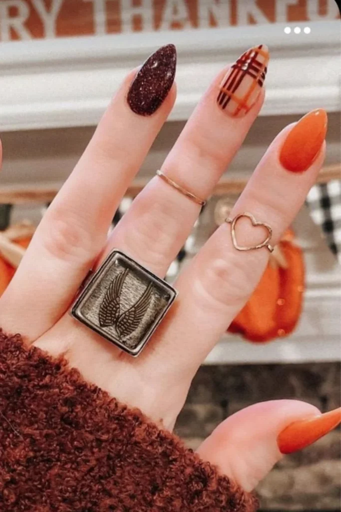 Autumnal September nails ideas featuring almond-shaped nails styled with a mix of burnt orange, deep maroon glitter, and a nude accent nail decorated with a red and orange plaid pattern